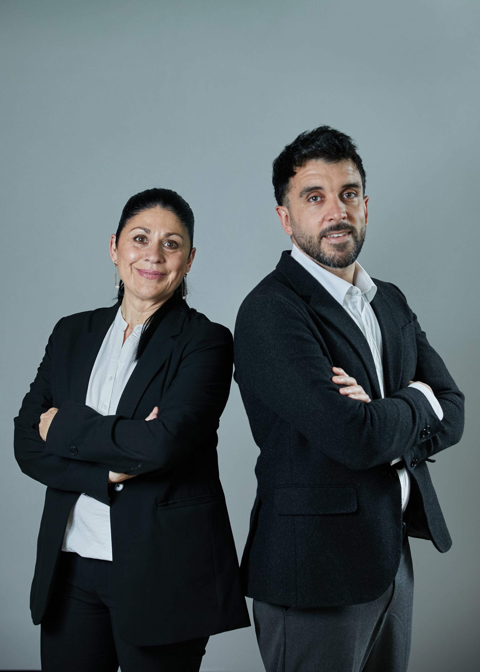 Retrato corporativo de dos profesionales —una mujer y un hombre— vestidos con traje oscuro y camisa blanca, de pie espalda con espalda y con los brazos cruzados, mirando a cámara sobre fondo neutro gris.