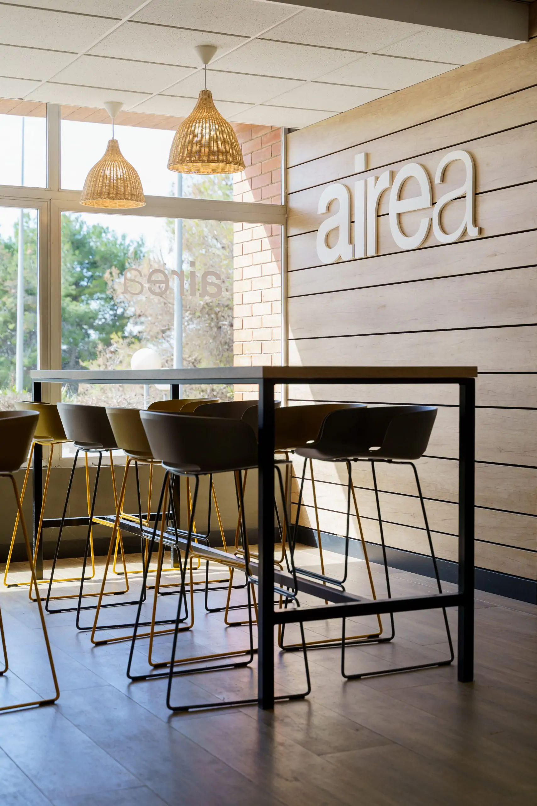 Zona de mesas altas y taburetes bajo el rótulo “Airea” junto a grandes ventanales.