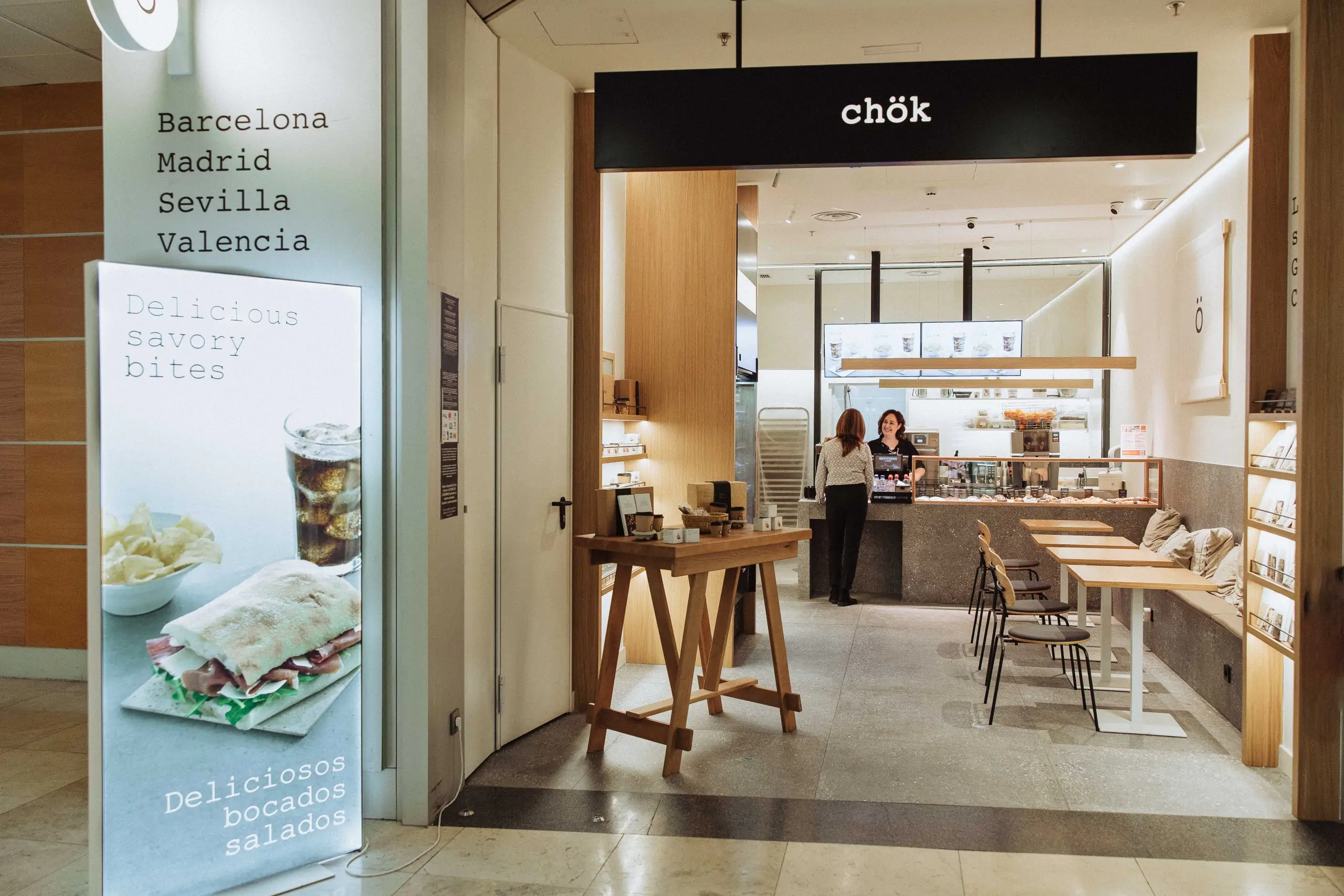 Entrada de cafetería en centro comercial con mostrador y zona de mesas en el interior.