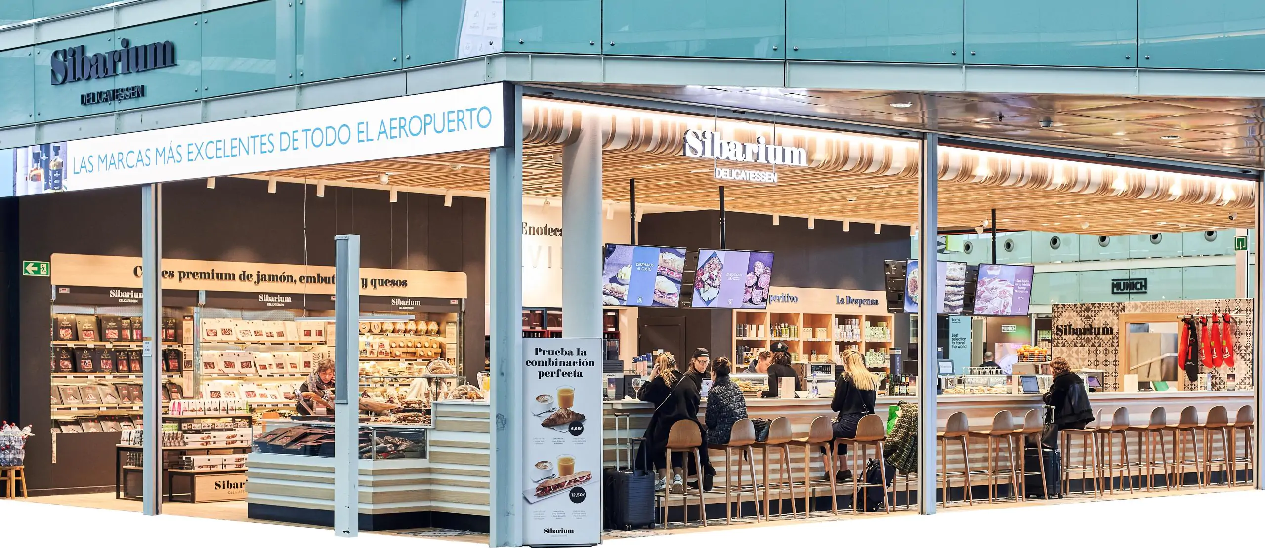 Local de delicatessen en aeropuerto con barra, vitrinas de productos y clientes sentados.