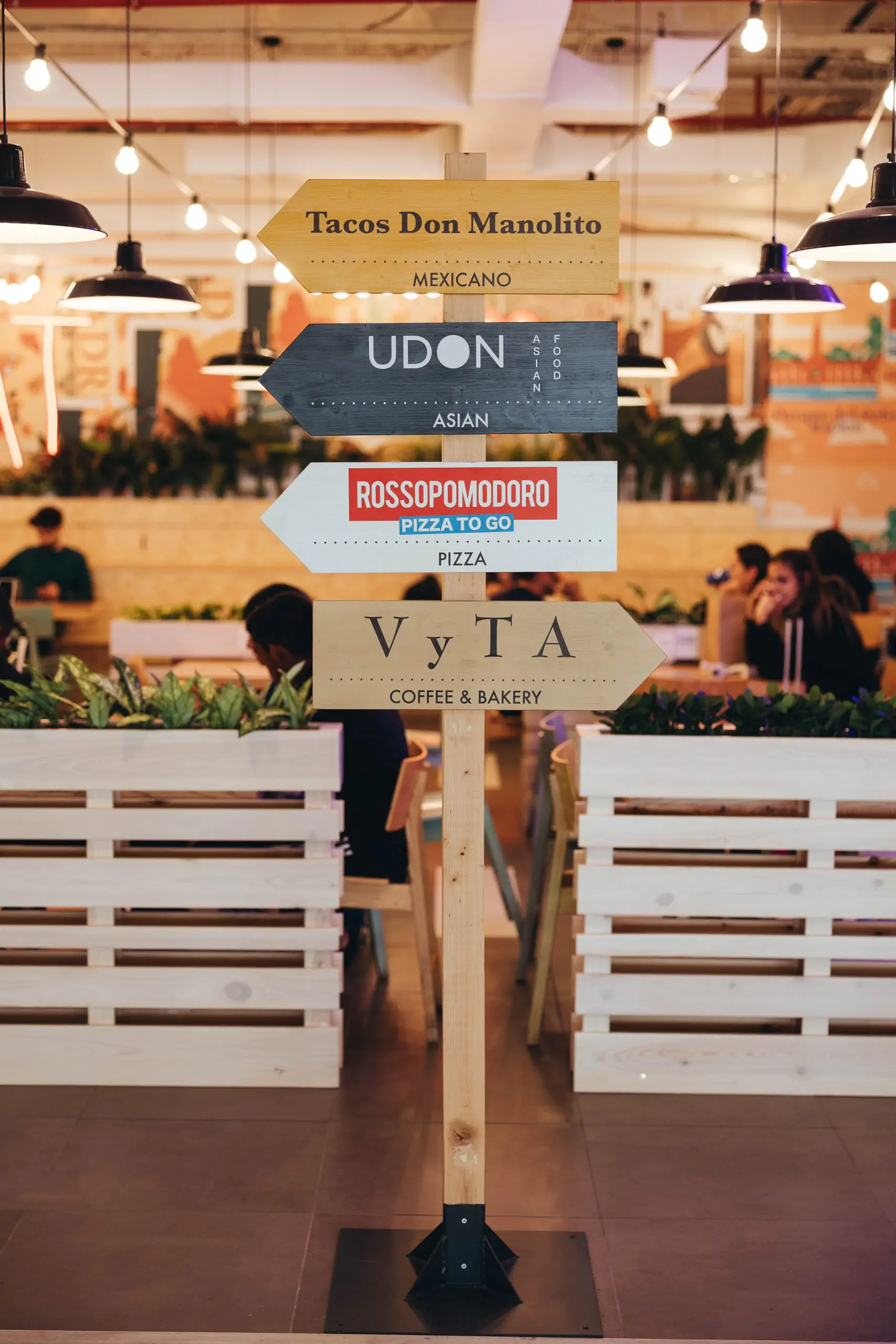 Señal direccional con nombres de restaurantes en zona de restauración interior.