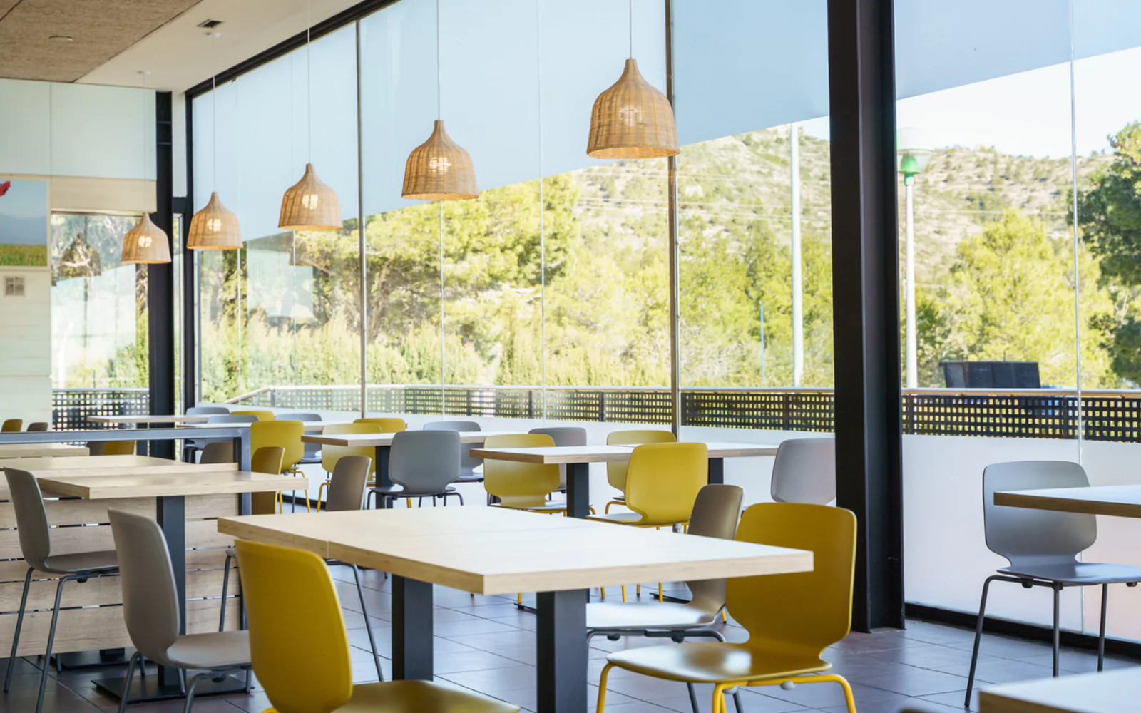 Comedor luminoso con mesas y sillas amarillas junto a grandes ventanales con vistas al exterior.