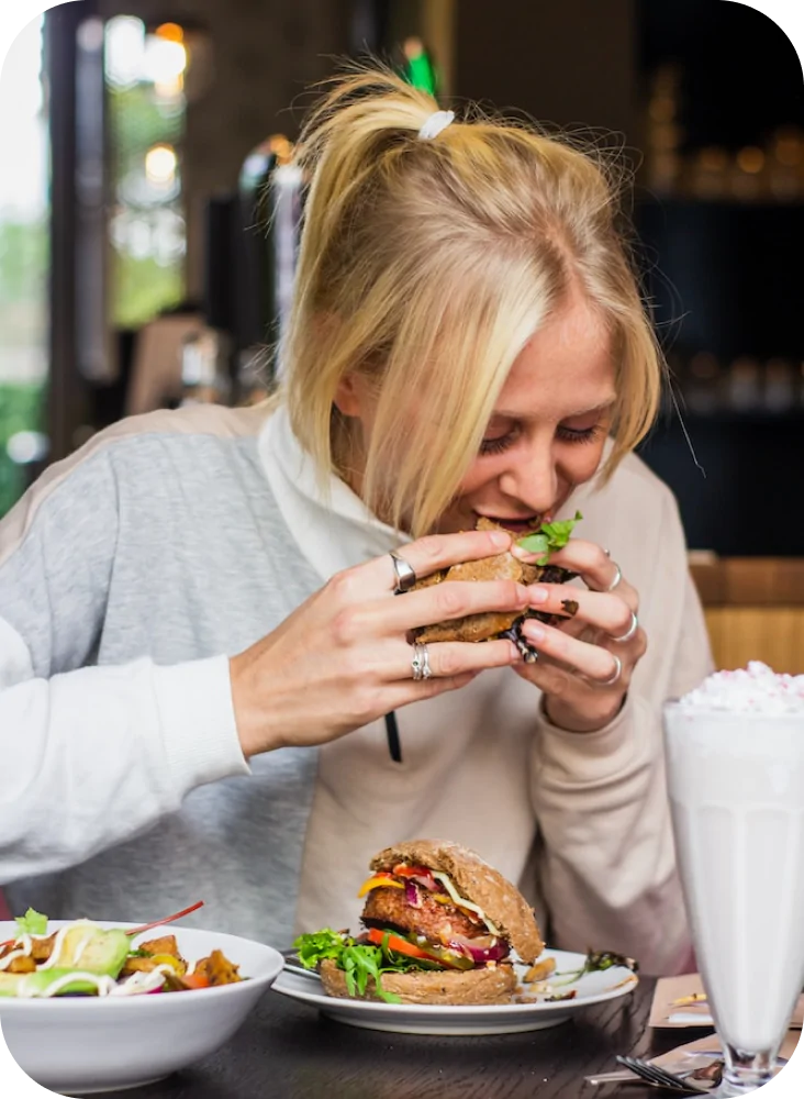 Mujer comiendo una hamburguesa en restaurante con plato y batido sobre la mesa.