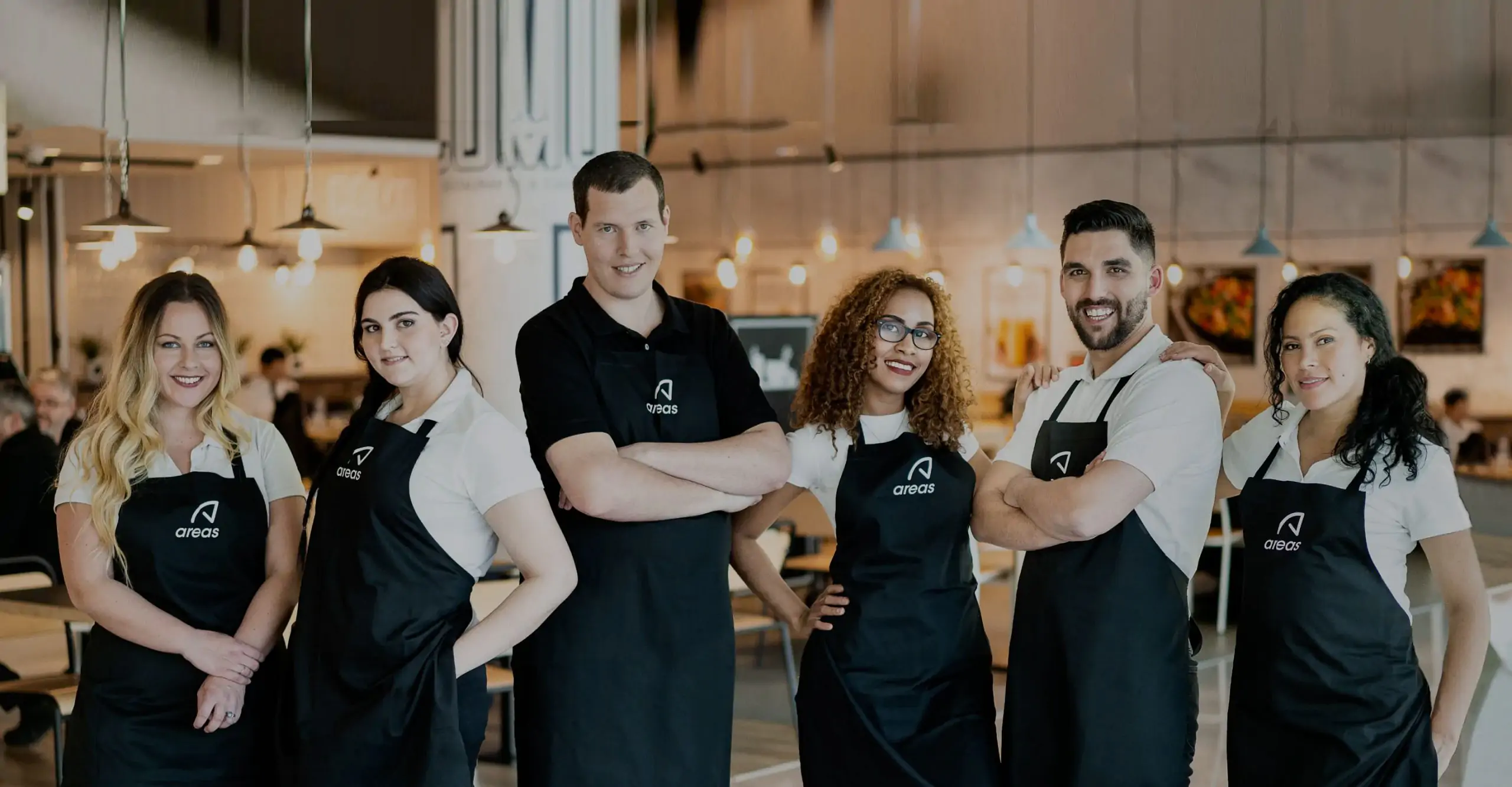 Equipo de seis empleados de restauración posando en el interior de un local, vestidos con polo blanco y delantal negro con el logotipo “areas”, de pie en fila y sonriendo a cámara, con el restaurante iluminado al fondo.
